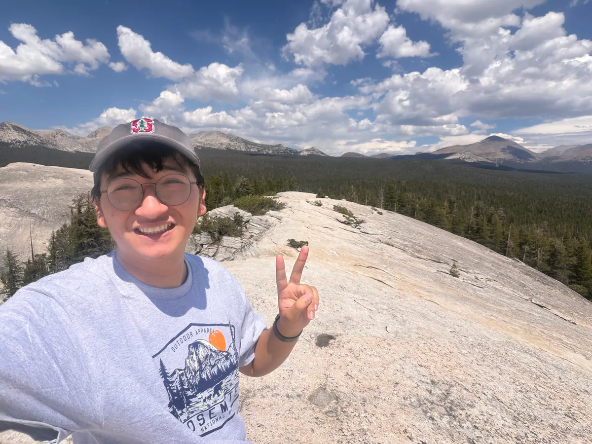 Krish Wu smiling on a bare granite dome, with pine forest and mountains behind him